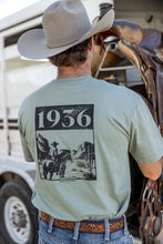Load image into Gallery viewer, Man wearing a light green t-shirt with a graphic and text, standing next to a horse trailer with a saddle.